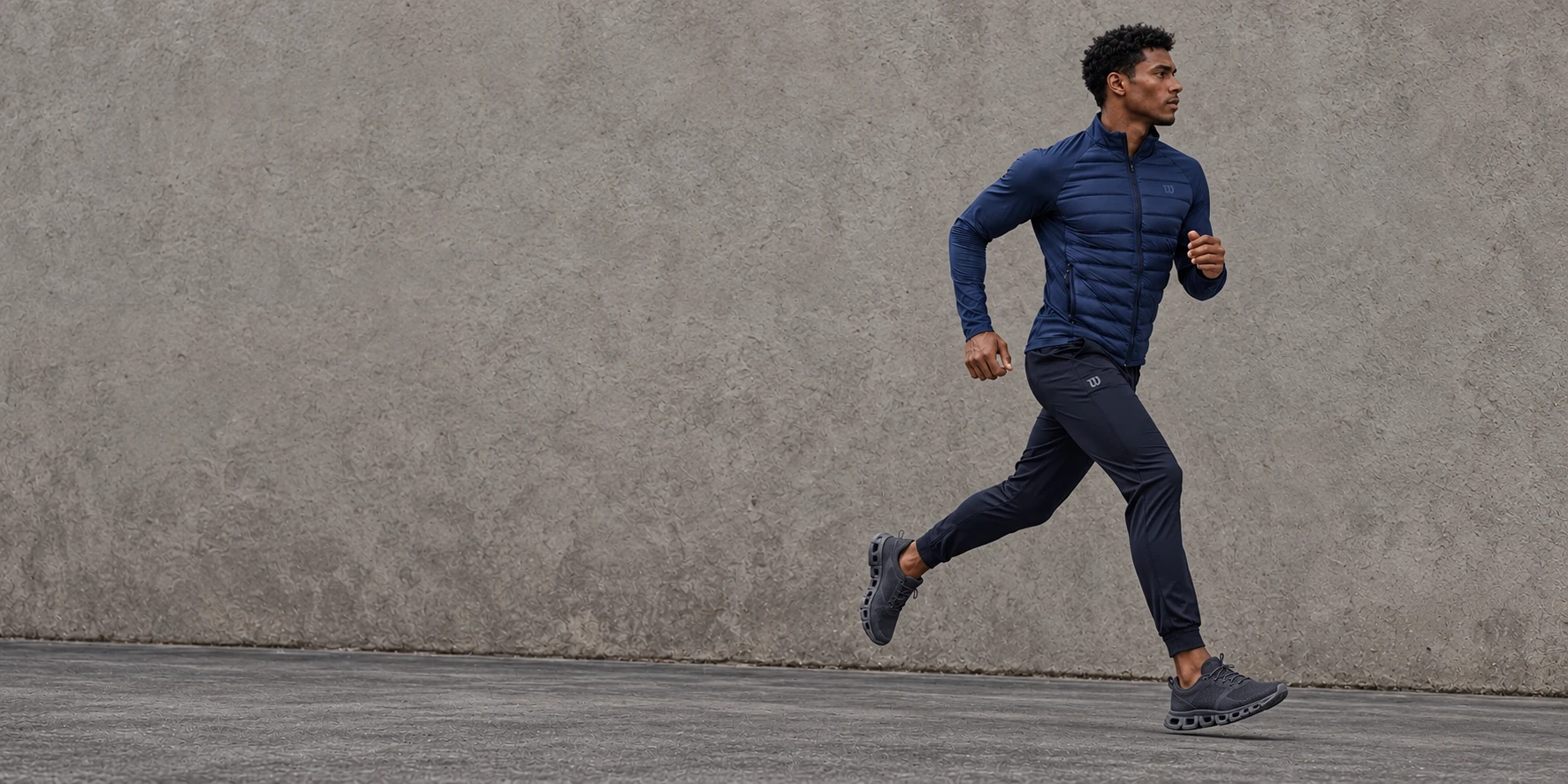 Atleta corriendo con chamarra azul marino, pants oscuro y tenis gris carbón frente a un muro gris piedra